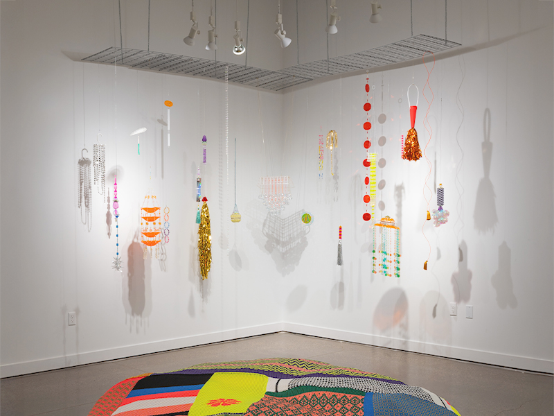 Installation view of a corner gallery with multiple vibrant, handcrafted hanging sculptures made from beads, plastic, tinsel and fringe. The ornaments are hanging from the ceiling, casting soft shadows on the white walls. A multicoloured woven mat with graphic patterns is on the floor.