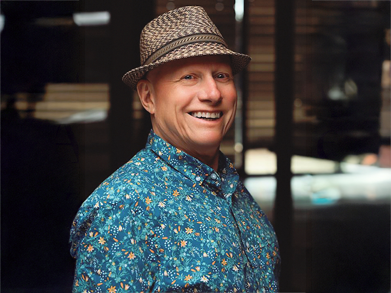 Wes Jenkins in a blue patterned shirt, fedora hat, standing on an angle looking forward and smiling
