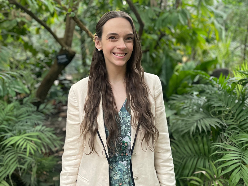 Caitlin Russell with long brown hair standing against green foliage wearing a cream-coloured blazer