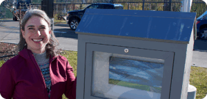 NEMG Amada Reber gallery Artist Amanda Reber stands beside an outdoor mini gallery displaying their artwork. The artist has long hair, glasses, and is wearing a red zippered jacket over a light brown sweater. There is grass and a street in the background.