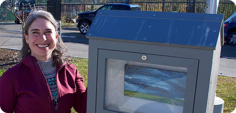 Artist Amanda Reber stands beside an outdoor mini gallery displaying their artwork. The artist has long hair, glasses, and is wearing a red zippered jacket over a light brown sweater. There is grass and a street in the background.