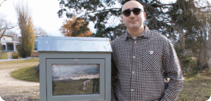 NEMG Amir Salehi gallery Artist Amir Salehi stands beside an outdoor mini gallery displaying their artwork. The artist is wearing sunglasses and a checked button-up shirt, and there are trees in the background.