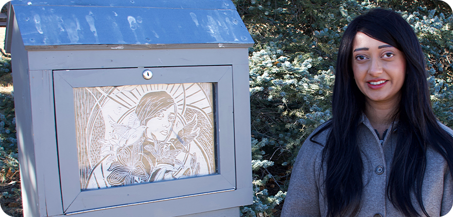 Artist Bukhtawar Malik stands beside an outdoor mini gallery displaying their artwork. The artist has long dark hair and is wearing a grey button-up coat, and there is a tree in the background.