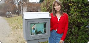 NEMG Christina Yao gallery Artist Christina Yao stands beside an outdoor mini gallery displaying their artwork. The artist has medium length hair and is wearing a red cardigan over a white tee, and jeans. There is a sidewalk, grass and shrubs in the background.