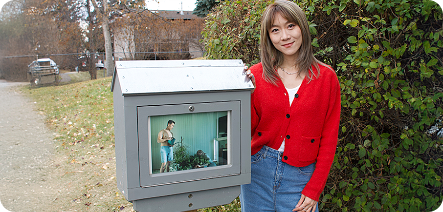 Artist Christina Yao stands beside an outdoor mini gallery displaying their artwork. The artist has medium length hair and is wearing a red cardigan over a white tee, and jeans. There is a sidewalk, grass and shrubs in the background.