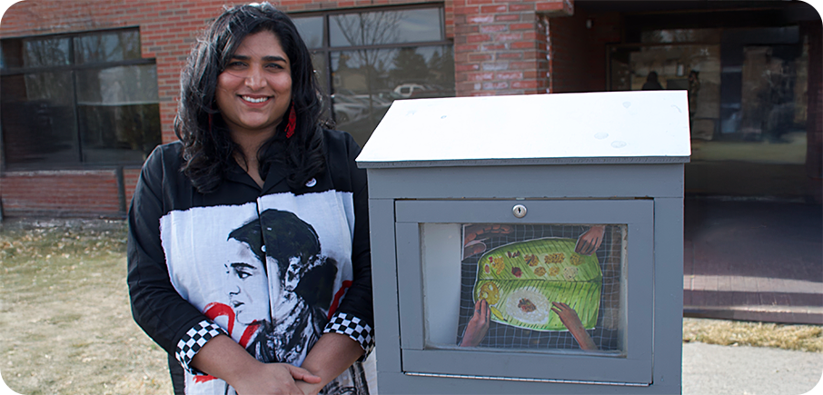 Artist Ciza Zoya stands beside an outdoor mini gallery displaying their artwork. The artist has long black hair and is wearing a black shirt with a black-and-white portrait of a woman on it. There is a building in the background.