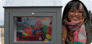 NEMG Justine Pozon gallery Artist Justine Pozon stands beside an outdoor mini gallery displaying their artwork. The artist has glasses and black hair with a white streak in the bangs, and is wearing a multicolour patterned scarf over a light brown jacket.
