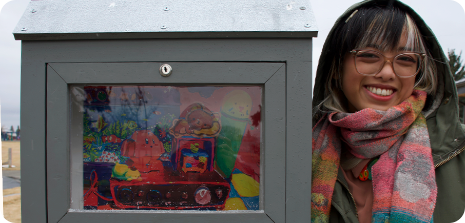 Artist Justine Pozon stands beside an outdoor mini gallery displaying their artwork. The artist has glasses and black hair with a white streak in the bangs, and is wearing a multicolour patterned scarf over a light brown jacket.