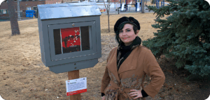 NEMG Mariella Villalobos gallery Artist Mariella Villalobos stands beside an outdoor mini gallery displaying their artwork. The artist has short dark hair and is wearing a black toque and turtleneck sweater, with a brown belted coat. There is a field of grass, an evergreen tree and a children's park in the background.