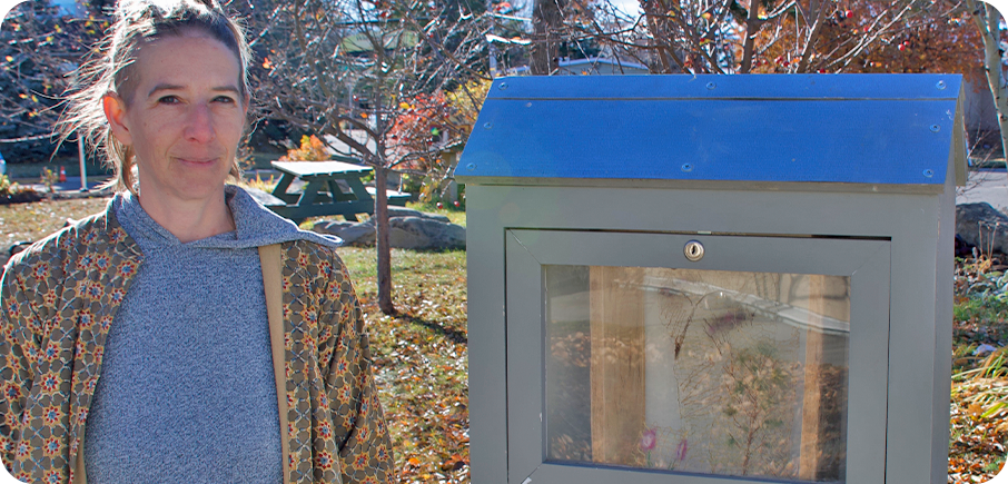 Artist Narda Ray stands beside an outdoor mini gallery displaying their artwork. The artist has light brown hair that is worn up, and is wearing a light blue hooded sweater under a brown patterned jacket. There is grass, trees and a picnic table in the background.