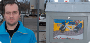 NEMG Paul Piotto gallery Artist Paul Piotto stands beside an outdoor mini gallery displaying their artwork. The artist has short brown hair dark shirt under a blue zippered jacket. There are some trees, driveways and cars in the background.