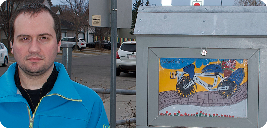 Artist Paul Piotto stands beside an outdoor mini gallery displaying their artwork. The artist has short brown hair dark shirt under a blue zippered jacket. There are some trees, driveways and cars in the background.