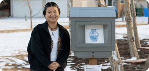 NEMG Rachel Hao Ran Li gallery Artist Rachel Hao Ran Li 黎浩然 stands beside an outdoor mini gallery displaying their artwork. The artist has short black hair pinned back with barrettes, and is wearing a dark jacket over a white shirt, as well as earrings and necklaces. There is snow on the ground, as well as some trees and houses in the background.