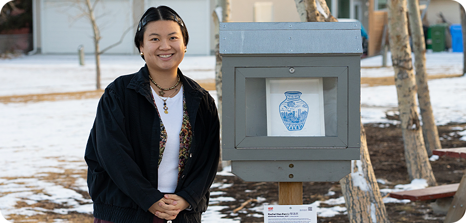 Artist Rachel Hao Ran Li 黎浩然 stands beside an outdoor mini gallery displaying their artwork. The artist has short black hair pinned back with barrettes, and is wearing a dark jacket over a white shirt, as well as earrings and necklaces. There is snow on the ground, as well as some trees and houses in the background.