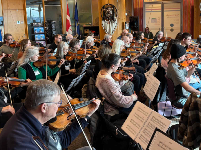A room of people sitting in front of music stands playing fiddles. On the wall is an Indigenous textile artwork and the Alberta and Canada flags stand next to it.