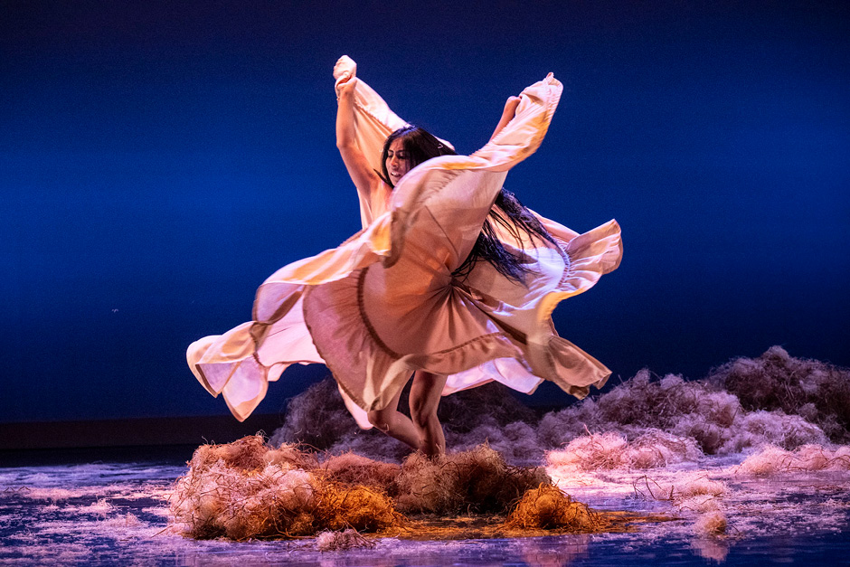 a dancer with long dark hair and billowing, flowing dress appears to be twirling on stage