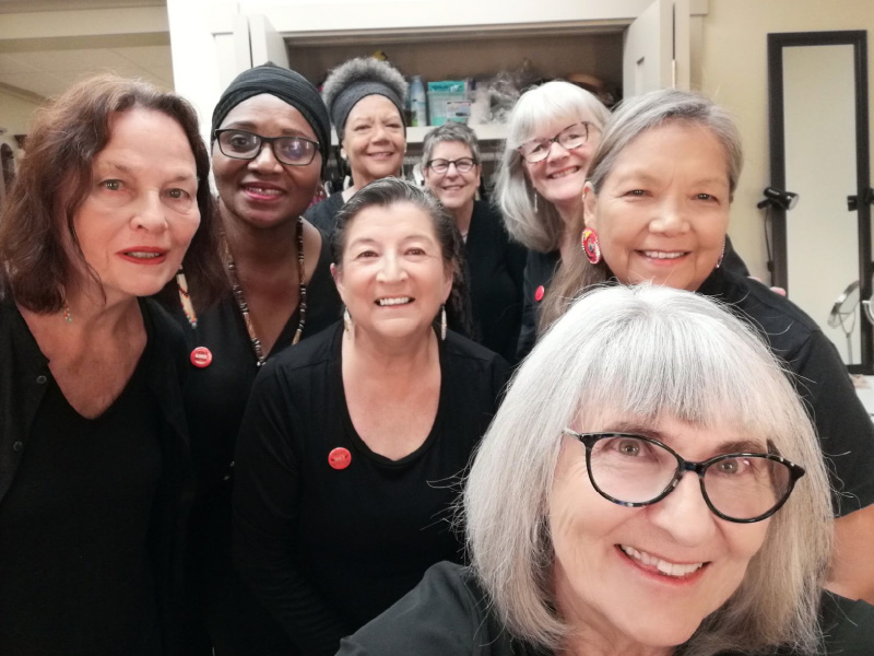 Eight women crowd together, all wearing dark clothing, smiling and facing forward in a small room