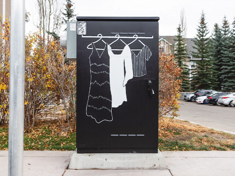 A utility box painted black with simple white outlines of three pieces of clothing hanging on a rod.