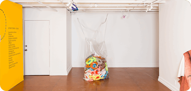 A clear plastic bag filled with colourful soft sculptures sits in the middle of a white gallery space with a yellow wall panel on the left.