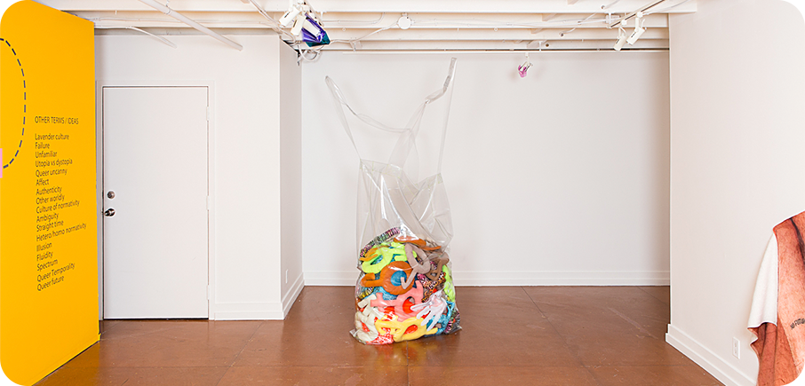 A clear plastic bag filled with colourful soft sculptures sits in the middle of a white gallery space with a yellow wall panel on the left.