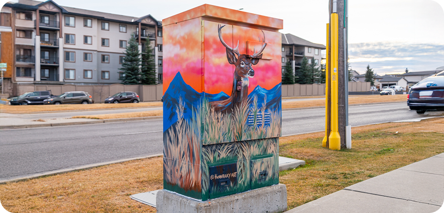 A utility box painted with a deer standing in tall grass against blue mountains and a bright pink-orange sky.