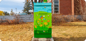 A utility box painted with a bright green field of yellow flowers, bees, and a person lying in the grass reading under a blue sky with clouds.