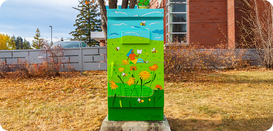 A utility box painted with a bright green field of yellow flowers, bees, and a person lying in the grass reading under a blue sky with clouds.