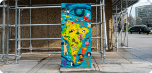 A brightly painted utility box showing a colourful cartoon-style map of South America, filled with animals, people, foods and cultural symbols, with a large blue eye at the top.