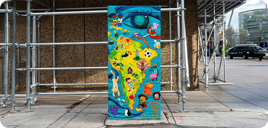 A brightly painted utility box showing a colourful cartoon-style map of South America, filled with animals, people, foods and cultural symbols, with a large blue eye at the top.