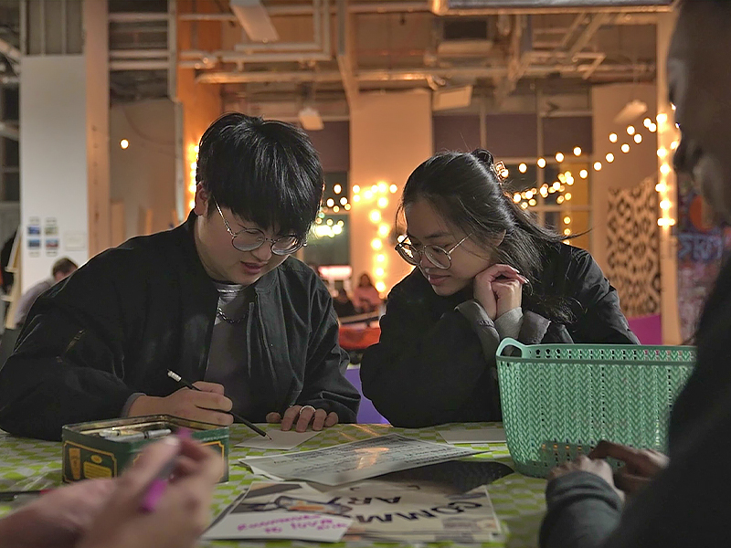 Two people sitting side-by-side, one drawing and the other looking at what is being drawn. There are drawings all over the table they are at and there are hanging lights behind them.