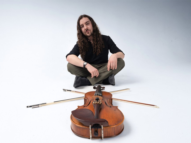 James Watson sits against a white background wearing dark clothes. He is cross-legged with hands on his knees, a violin and two bows are in front of him.