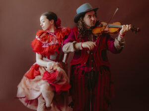 James Watson plays a violin while wearing a hat and red suit. He is standing next to a woman looking to the side wearing a red and white flamenco outfit