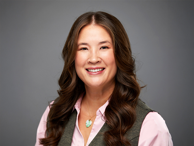 Kim Tyers smiling, facing forward, shown from the shoulders up with long brown hair against a grey background.