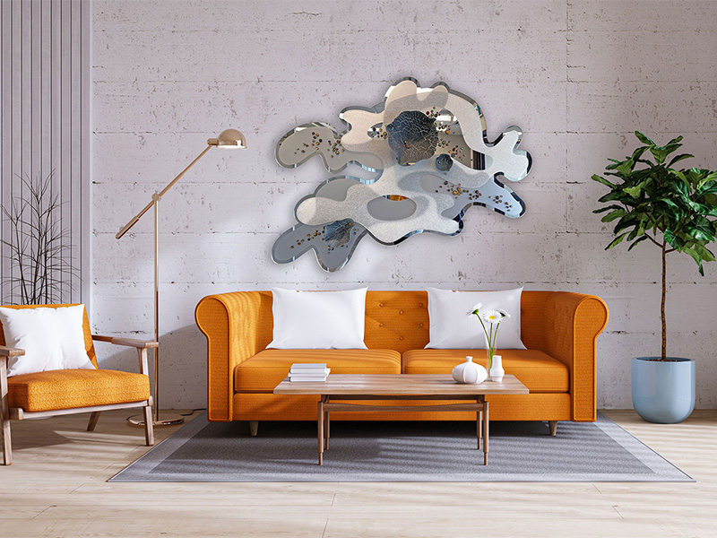A room with white wall, a large glass artwork is on the wall which looks like a blob. There is an orange couch with two white pillows, a coffee table and a stand-up lamp.