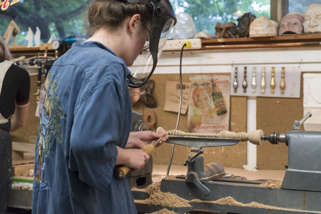 A worker with protective clothing and mask working on a lathe machine with what looks like a wood spindle, in a workshop