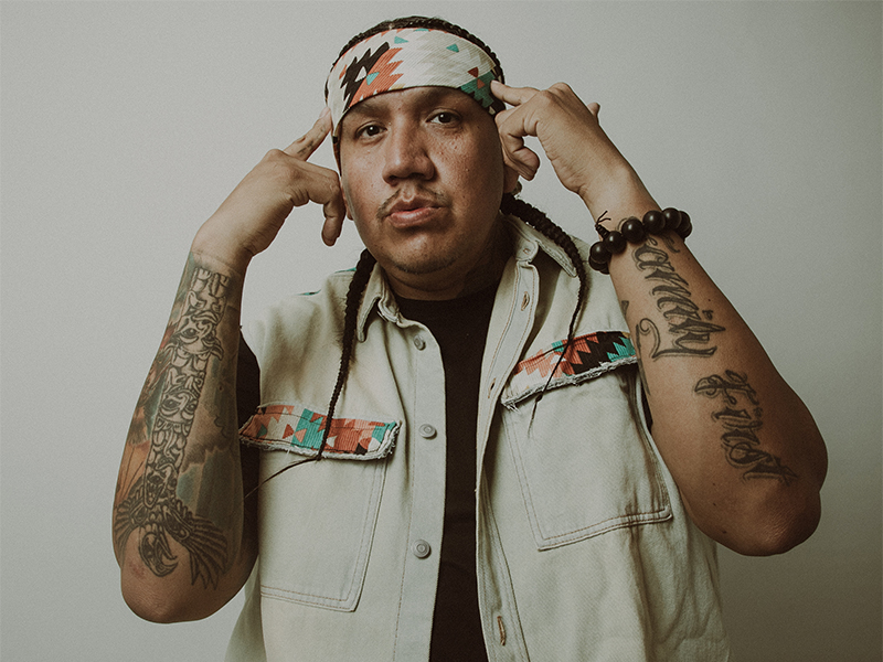 Drezus stands with both hands held up to his head, fingers on temples. He is wearing a beige vest and head band, his arms are tattooed.