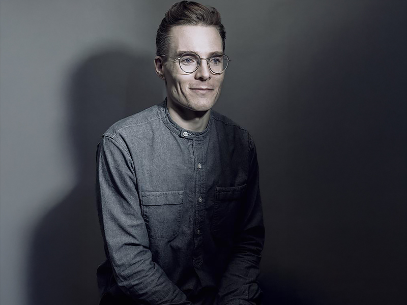 Tyler Los-Jones wearing a grey, long-sleeved shirt sitting in a darkened room with what appears to be a small light shining on his face and right side of his upper body.