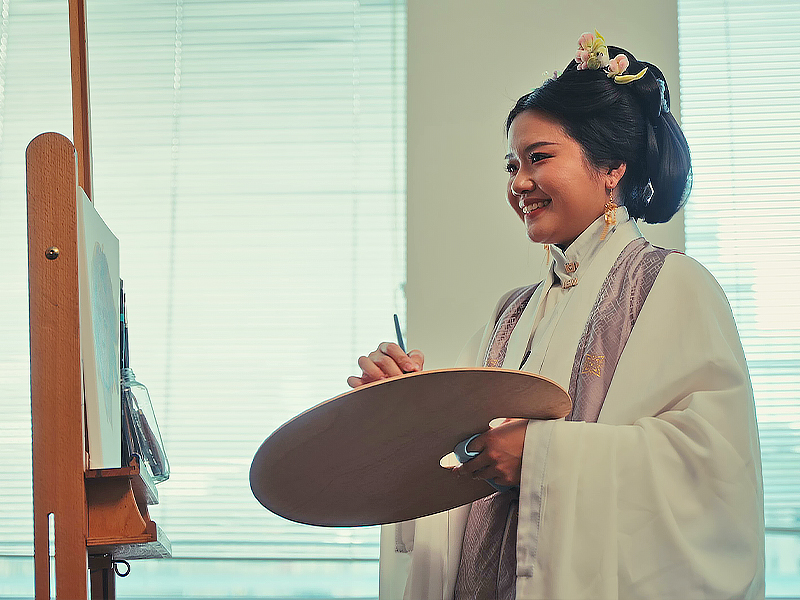 Artist Yu Chen in a studio standing at an art easel hosing a palette and paintbrush, while wearing traditional Chinese clothing and a floral headpiece.